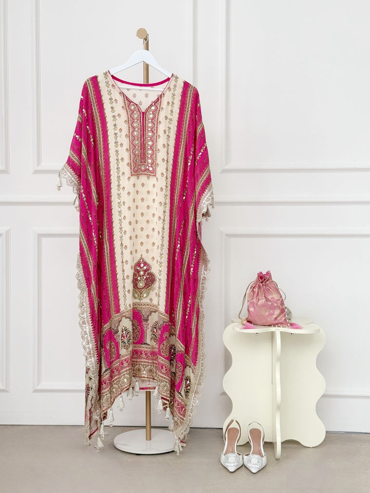 The ivory central panel features elegant floral motifs, beautifully framed by deep pink side panels with vertical gold accents. The neckline showcases an intricately embroidered V-shaped design with mirror work and metallic thread detailing, adding a touch of regal flair. Wide, flowy sleeves with lace trim and tassels enhance the kaftan’s graceful silhouette, while the hemline boasts rich embroidery and tassel accents for a stunning finish.