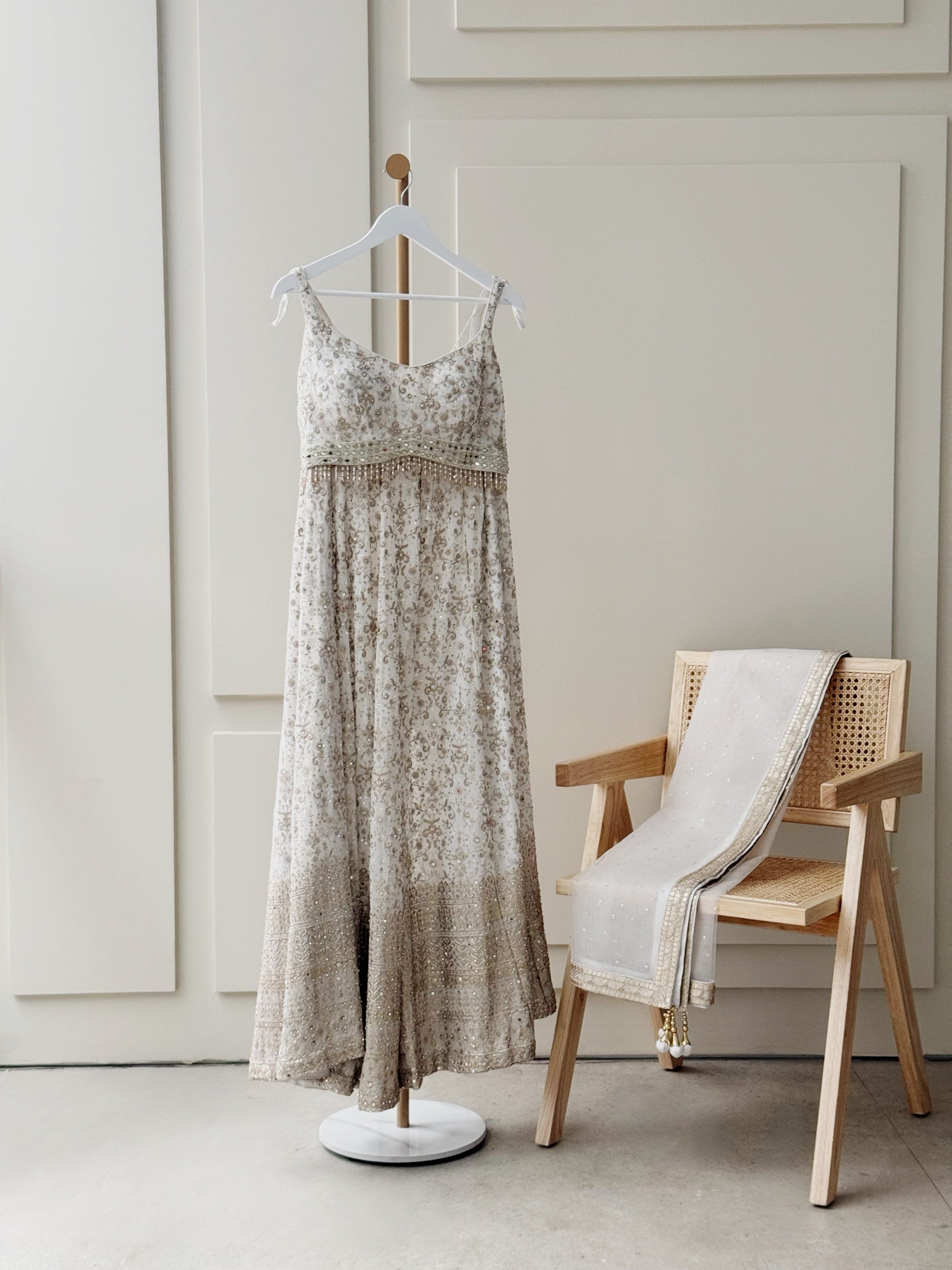 An elegant embroidered sleeveless dress in muted gold and beige tones hangs on a white hanger, featuring a delicate V-neckline, thin tie-up straps, and an embellished waistband. The flowing skirt is adorned with intricate floral and paisley embroidery, with a heavily embellished hemline. Beside the dress, a wooden chair with a woven cane seat and backrest holds a matching embroidered dupatta with gold detailing and tassels. The setup is placed against light-paneled walls, creating a minimal and sophisticate