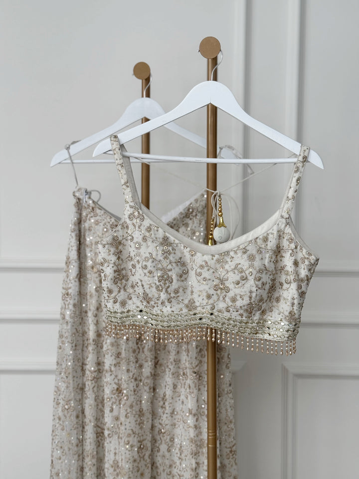 An elegant embroidered sleeveless dress in muted gold and beige tones hangs on a white hanger, featuring a delicate V-neckline, thin tie-up straps, and an embellished waistband. The flowing skirt is adorned with intricate floral and paisley embroidery, with a heavily embellished hemline.