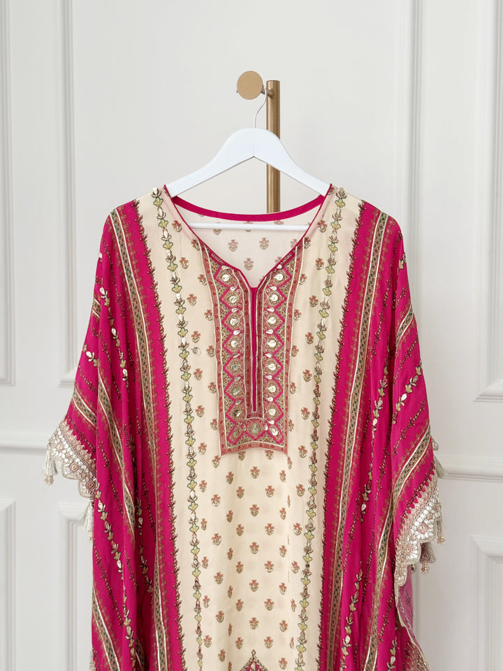 The ivory central panel features elegant floral motifs, beautifully framed by deep pink side panels with vertical gold accents. The neckline showcases an intricately embroidered V-shaped design with mirror work and metallic thread detailing, adding a touch of regal flair. Wide, flowy sleeves with lace trim and tassels enhance the kaftan’s graceful silhouette, while the hemline boasts rich embroidery and tassel accents for a stunning finish.