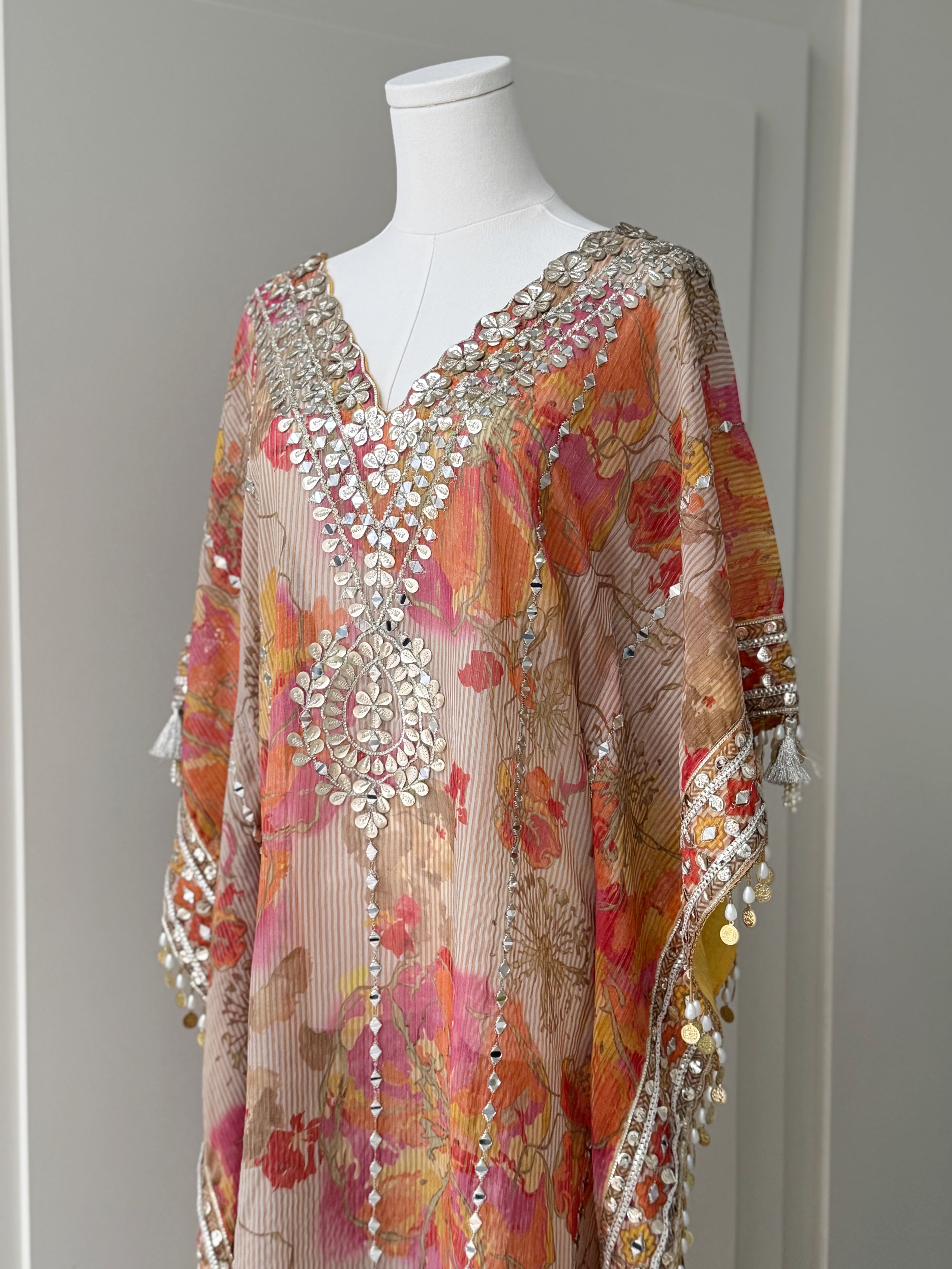 Floral printed kaftan in orange, pink, and yellow hues with intricate mirror work on the neckline and borders, finished with tassel details, ideal for festive and casual occasions