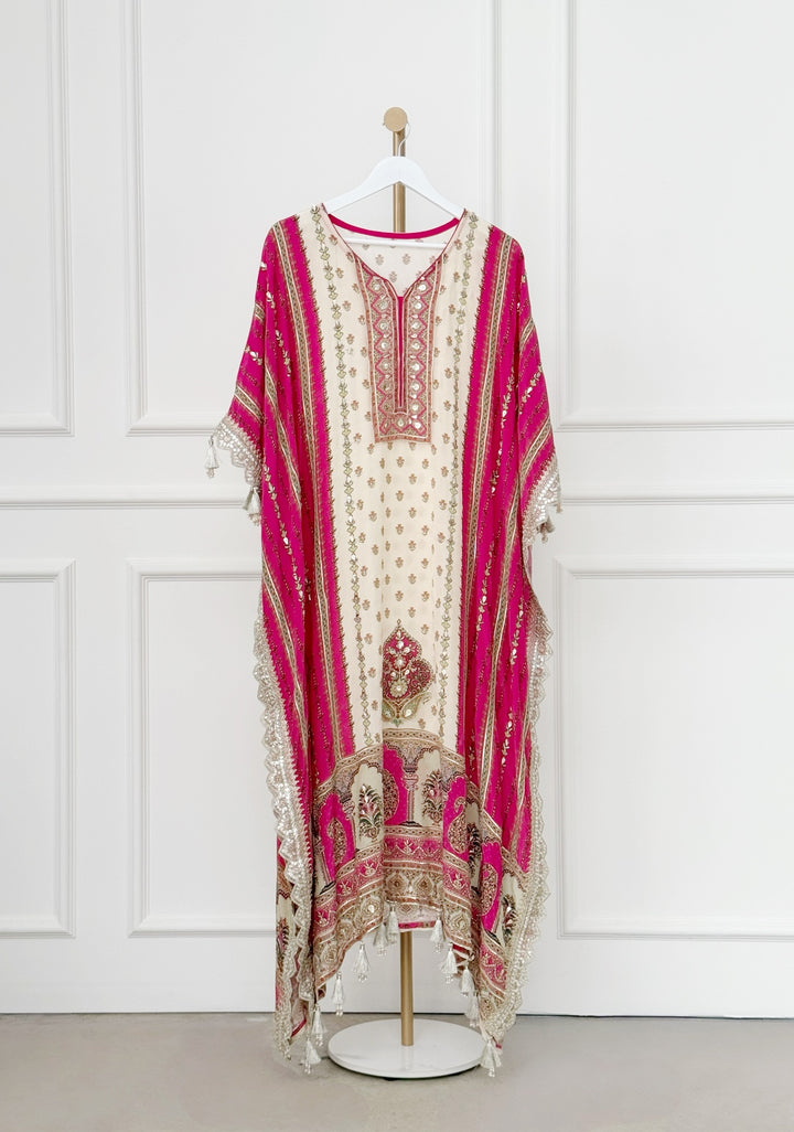 The ivory central panel features elegant floral motifs, beautifully framed by deep pink side panels with vertical gold accents. The neckline showcases an intricately embroidered V-shaped design with mirror work and metallic thread detailing, adding a touch of regal flair. Wide, flowy sleeves with lace trim and tassels enhance the kaftan’s graceful silhouette, while the hemline boasts rich embroidery and tassel accents for a stunning finish.