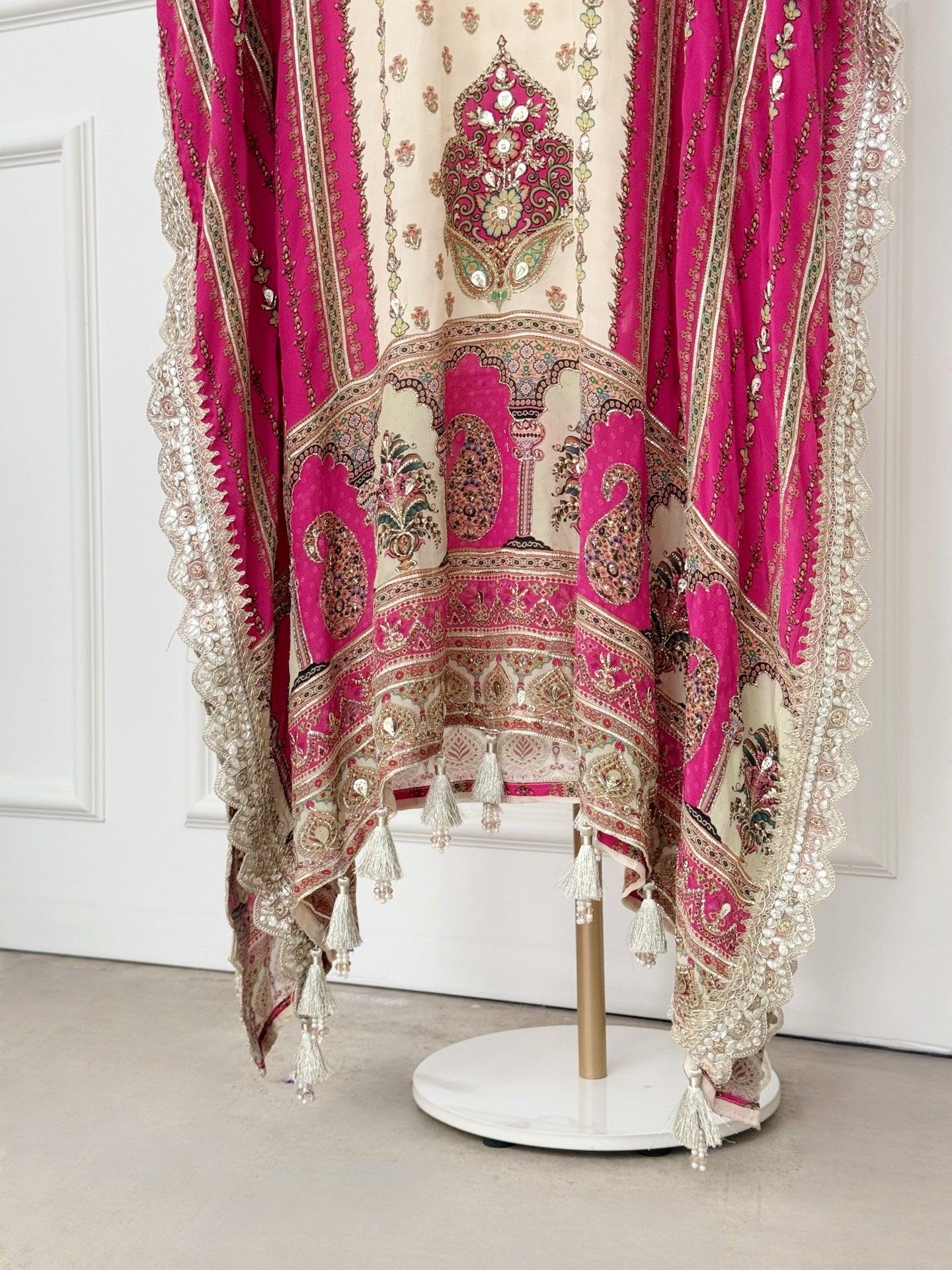 The ivory central panel features elegant floral motifs, beautifully framed by deep pink side panels with vertical gold accents. The neckline showcases an intricately embroidered V-shaped design with mirror work and metallic thread detailing, adding a touch of regal flair. Wide, flowy sleeves with lace trim and tassels enhance the kaftan’s graceful silhouette, while the hemline boasts rich embroidery and tassel accents for a stunning finish.